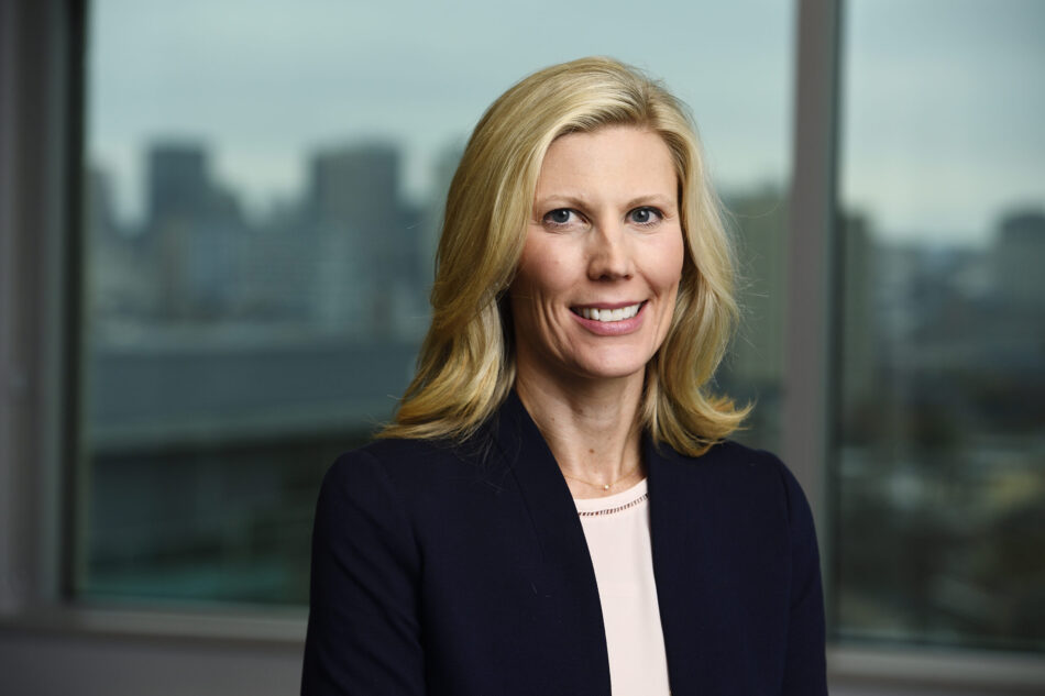 A headshot of Dr. Swenor is pictured in front of a window with a cityscape background. She is smiling warmly and wearing a navy-blue blazer with a light pink blouse and small gold and silver necklace.  