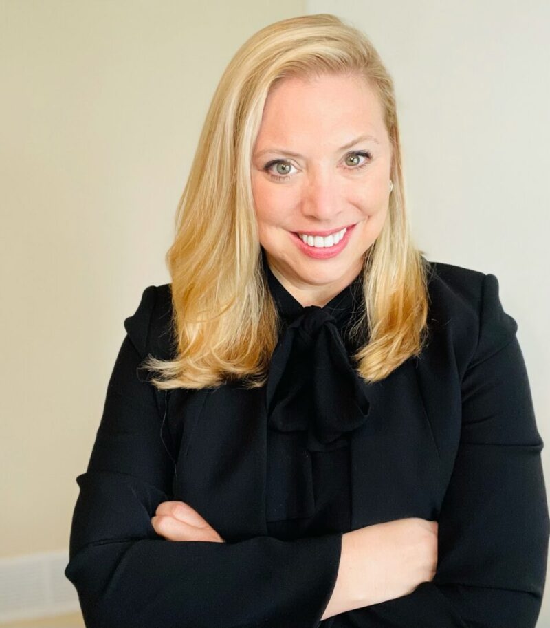 A headshot of Dr. Meeks is pictured in front of an ivory-colored background. She is smiling brightly and wearing a black blouse with a fashionable tie-knot at the front, with her arms crossed at the center.